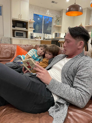 A man in a gray cardigan and black pants is reading a children’s book with two young boys on a brown leather couch.
