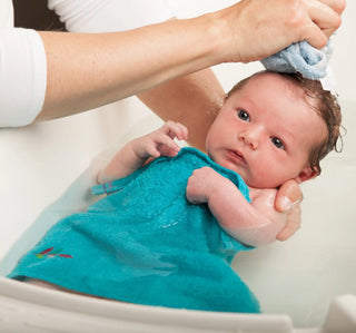 Baby wearing a turquoise outfit being bathed in water.