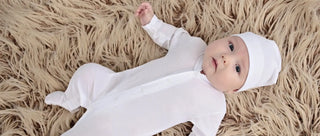 A baby wearing white clothing and a white hat lying on a furry surface.